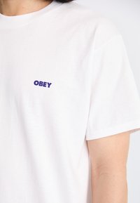 Cilvēks valkā baltu īsām piedurknēm t-kreklu ar mazu purpura "OBEY" logo kreisajā augšējā krūtī.