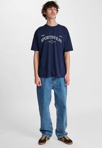 Marinblå bomulls-T-shirt med vit text och logodetaljer "SPORTSWEAR". Kombinerad med ljusblå jeans i denim och bruna sneakers.