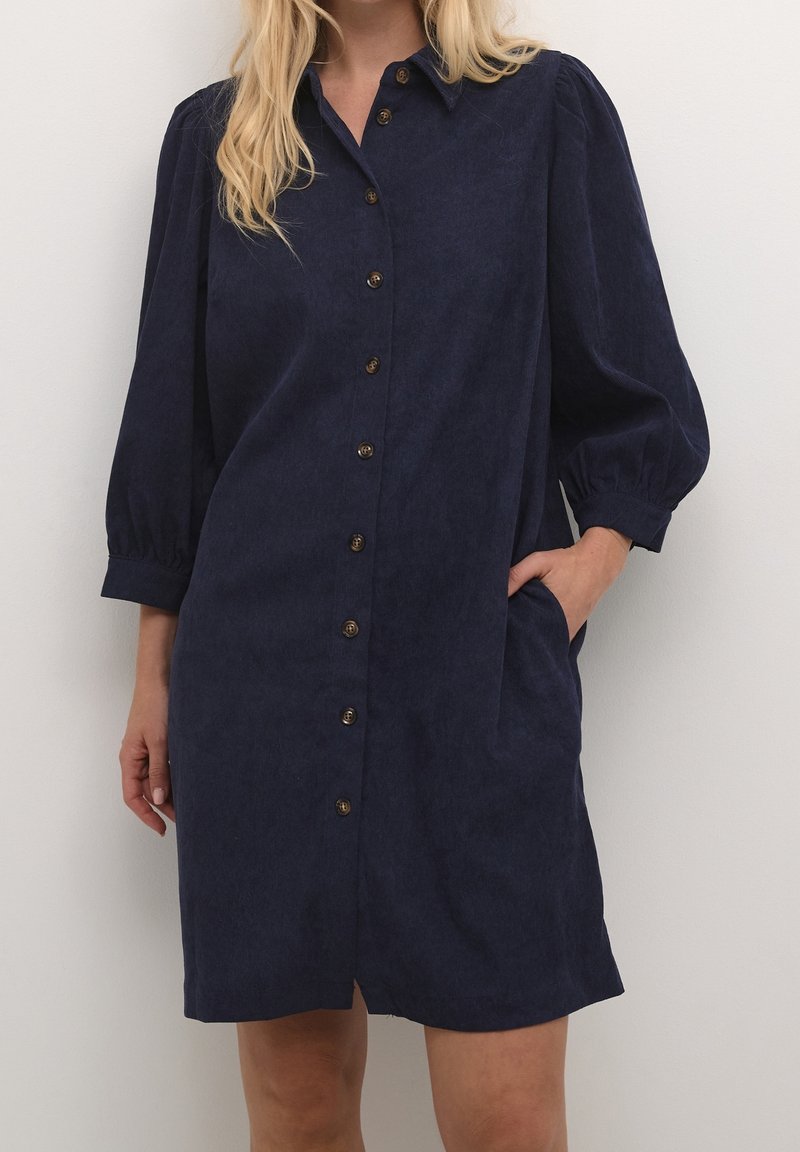 Femme portant une robe bleu marine boutonnée avec des manches bouffantes trois-quarts et une main dans une poche latérale, debout devant un fond uni.