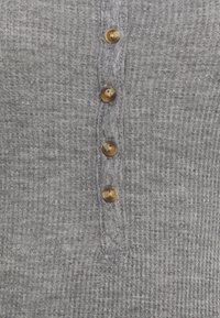 Grå stickad topp med ribbad textur, som har fyra runda, gyllene knappar längs en central knäppning. Mjuk tyg med en subtil lyster.