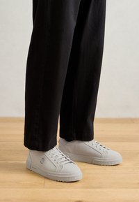 Baskets gris clair avec une finition texturée, bout rond et semelle plate, associées à un pantalon noir légèrement ample. Fond de sol en bois.