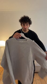 Sweatshirt cinza claro com mangas raglan, feito de tecido suave, apresentando um decote redondo, pendurado num cabide branco.