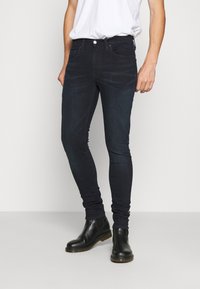 Tunna mörka denimjeans med en lätt glans, som har fem fickor, dragkedja fram och en åtsittande ankel, matchat med svarta läderstövlar vid ankeln.
