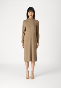 Abito in maglia marrone con maniche lunghe e colletto alto. Presenta slits laterali e una silhouette dritta. Indossato con sandali con tacco nudi.