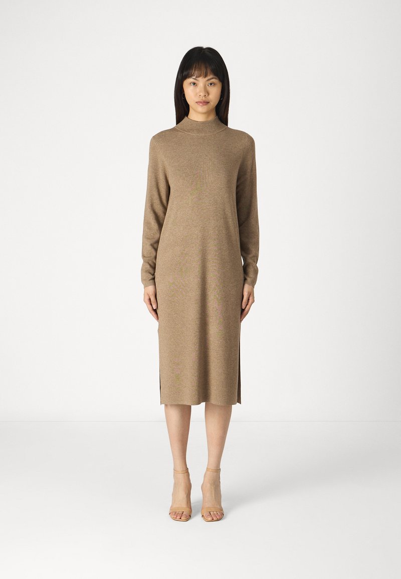 Abito in maglia marrone con maniche lunghe e colletto alto. Presenta slits laterali e una silhouette dritta. Indossato con sandali con tacco nudi.