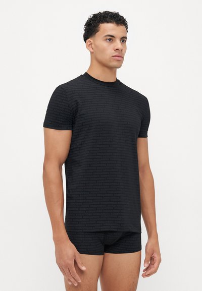 T-shirt nero a maniche corte con un motivo testurizzato che presenta il nome del marchio ripetuto. Vestibilità comoda e tessuto morbido. Indossato con slip neri coordinati.