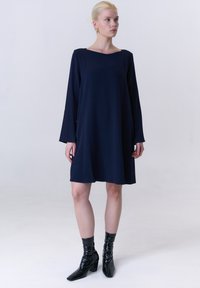 Navyblaues, langärmliges Kleid mit eckigem Ausschnitt, aus weichem Stoff gefertigt. Glatte Textur, knielang, kombiniert mit glänzenden schwarzen Stiefeletten.