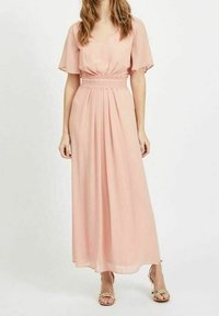 Robe maxi en chiffon rose doux avec un décolleté en V, des manches courtes évasées et une taille élastique ; jupe fluide avec des plis subtils ; portée avec des sandales dorées.