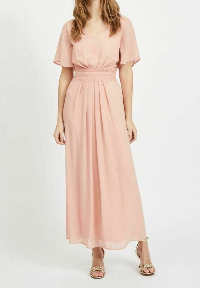 Robe maxi en chiffon rose doux avec un décolleté en V, des manches courtes évasées et une taille élastique ; jupe fluide avec des plis subtils ; portée avec des sandales dorées.