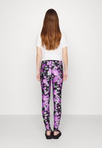 Leggings neri a fiori con grandi motivi di rose viola e verdi, tessuto liscia, forma aderente, indossati con una maglietta bianca e calzature nere.