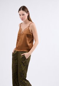 Camisón de satén marrón con encaje en el borde y tirantes ajustables, combinado con pantalones verdes con un estampado de manchas negras y naranjas.