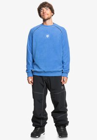Sweatshirt en polaire bleu avec des poignets côtelés, présente un logo brodé sur la poitrine. Assorti à un pantalon noir ample et des bottes noires.