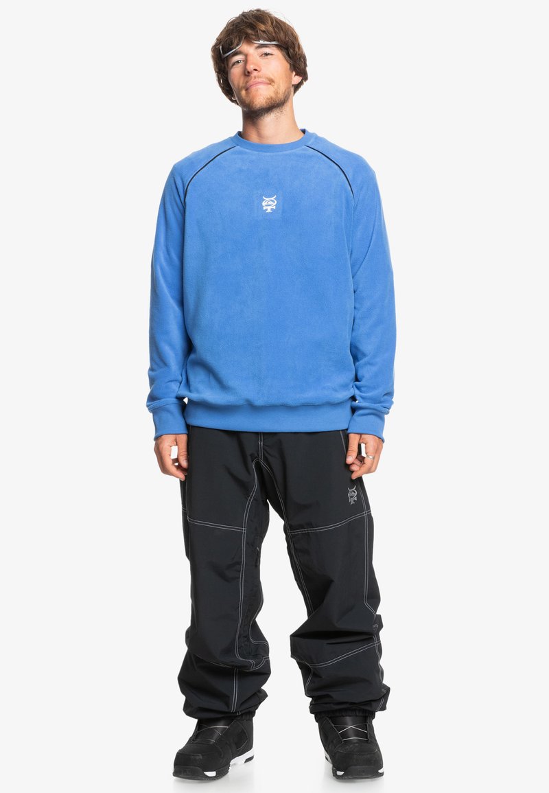 Blå fleece-tröja med ribbade manschetter, har en broderad logotyp på bröstet. Matchas med svarta, löst sittande byxor och svarta stövlar.