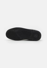 Semelle en caoutchouc noire avec des motifs de traction texturés, featuring un logo central et un design de talon galbé. Surface lisse avec des rainures définies.
