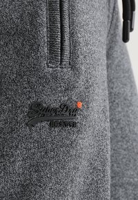 Sweat à capuche en tissu gris avec une finition texturée, doté d'une fermeture éclair au niveau du col et d'un patch logo noir avec des détails orange.
