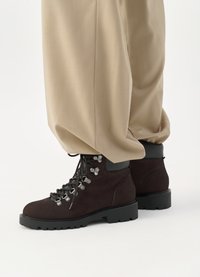 Brązowe zamszowe buty wędrówkowe z czarną gumową podeszwą, metalowymi oczkami i sznurówkami. Połączone z szerokimi beżowymi spodniami, ukazującymi dolną część stroju.