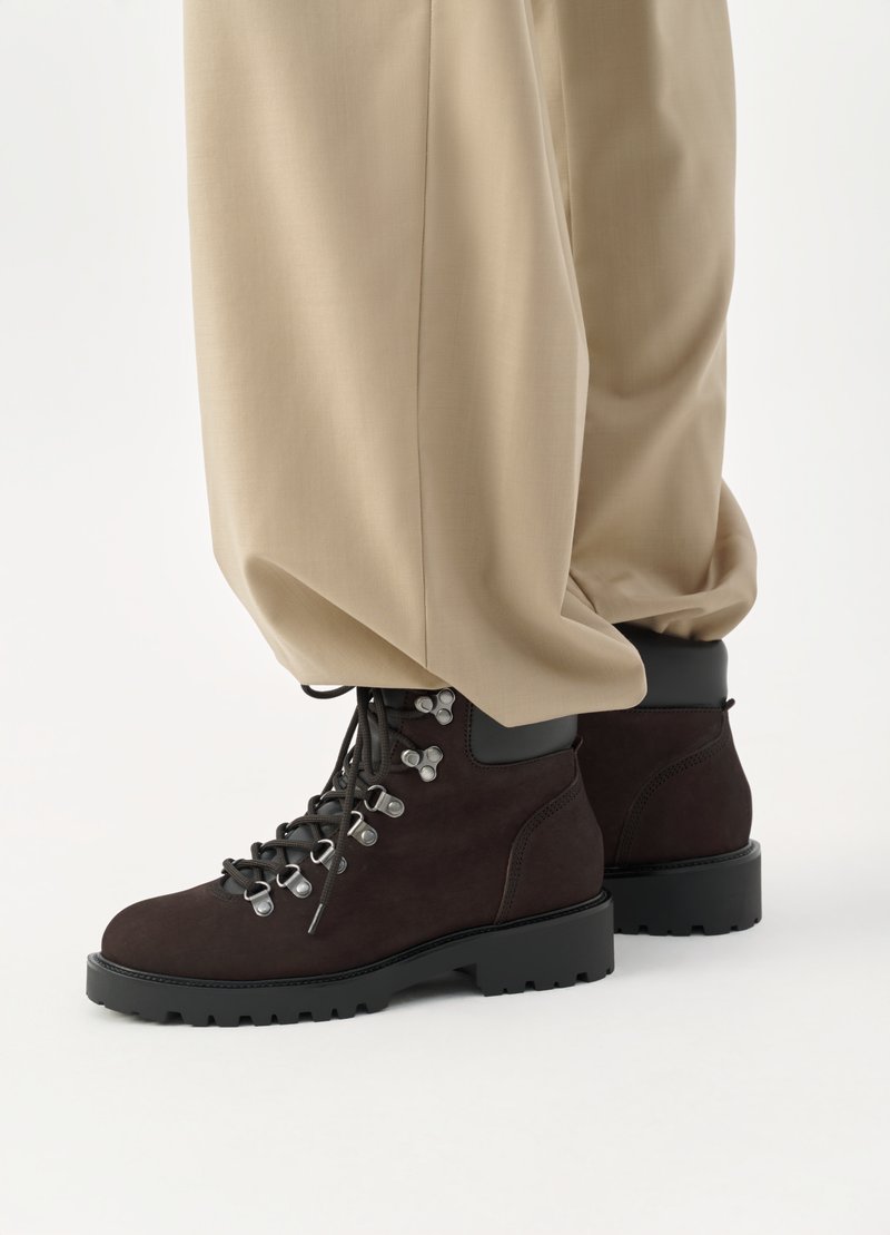 Bruine suède wandelschoenen met zwarte rubberen zolen, metalen oogjes en veters. Gecombineerd met brede beige broek, waarbij de onderste helft van de outfit zichtbaar is.