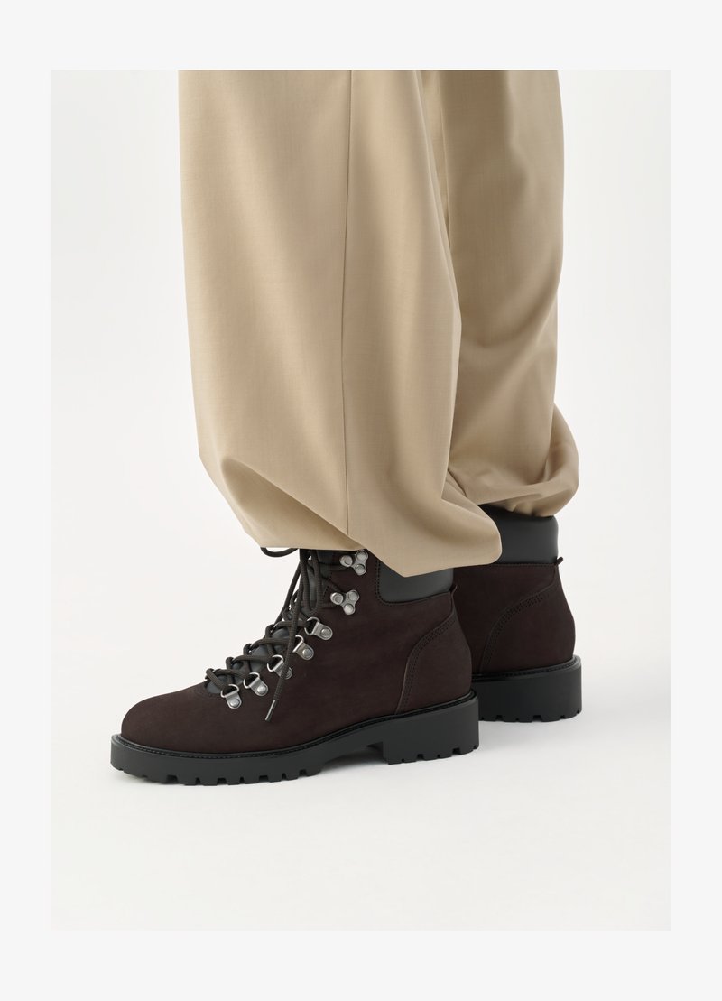 Bruine suède wandelschoenen met zwarte rubberen zolen, metalen oogjes en veters. Gecombineerd met brede beige broek, waarbij de onderste helft van de outfit zichtbaar is.