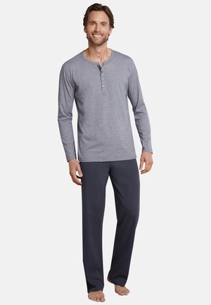 Leende man som står barfota iförd en grå långärmad henley-tröja och mörka pyjamasbyxor framför en enkel bakgrund.