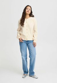 Cardigan crema con bottoni, texture morbida, scollatura rotonda, abbinato a jeans azzurri chiari e scarpe argentate, in piedi su uno sfondo neutro.