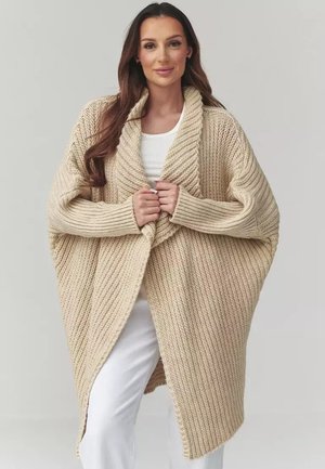 Kobieta w dużym beżowym dzianinowym kardiganie z kołnierzem szalowym, nałożonym na biały top i spodnie, stoi na prostym tle.