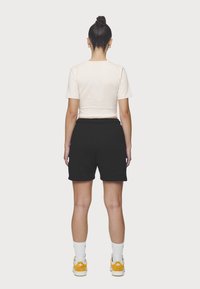 Černé šortky a světle béžové zkrácené tričko; modelka má na sobě bílé ponožky a žluté tenisky. Jednoduchý, neformální design s elastickým páskem.