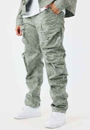 Hombre con pantalones cargo gris verdosos con múltiples bolsillos y zapatillas blancas, de pie con una mano en el bolsillo.