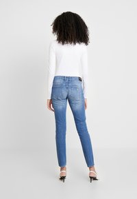 Pantalones vaqueros ajustados de mezclilla en color azul, con una cintura de altura media, bolsillos traseros y un lavado desgastado. Combinados con una camiseta blanca de manga larga.