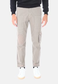 Pantaloni cargo beige con toppe al ginocchio, dettagli in cuciture rosse, tasche multiple, orli arrotolati e occhielli. Indossati con sneakers bianche.