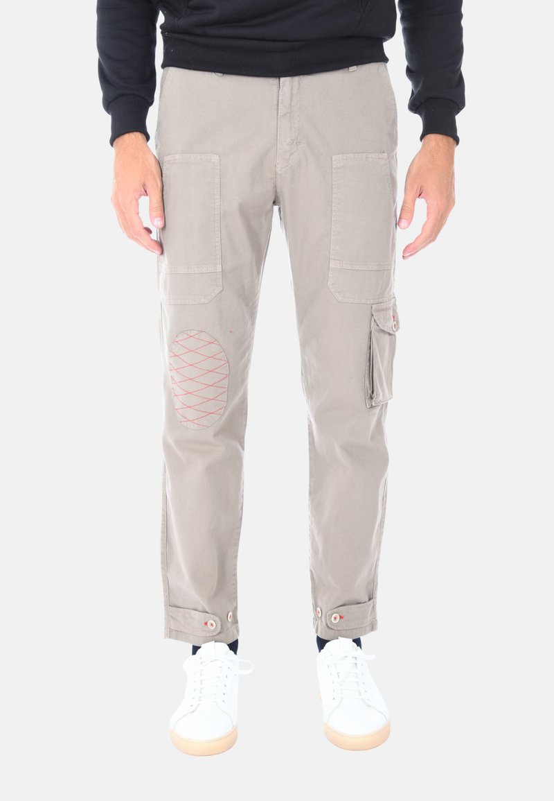 Pantaloni cargo beige con toppe al ginocchio, dettagli in cuciture rosse, tasche multiple, orli arrotolati e occhielli. Indossati con sneakers bianche.