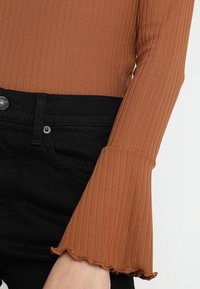 T-shirt à manches longues côtelé en orange brûlé, doté de manches évasées avec des bords en forme de scallop, associé à un jean noir taille haute.