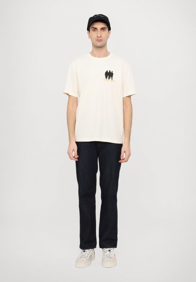 T-shirt crème à manches courtes avec un motif graphique noir, associé à un jean en denim sombre et des baskets blanches. Le modèle porte également une casquette noire.