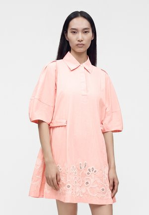 Femme aux longs cheveux noirs portant une robe rose ample à manches mi-longues avec une broderie florale blanche près de l'ourlet.
