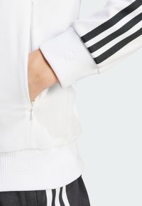 White athletic jacket with black three-stripe accents, ribbed cuffs, and a side zippered pocket. Smooth texture and fitted design.