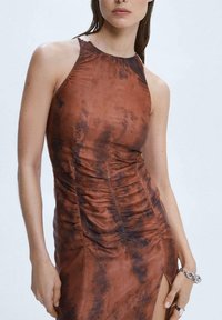 Frau in braunem, ärmellosem, hochgeschnittenem Kleid mit dunklem Batikmuster und seitlichem Schlitz, mit minimalem Schmuck am rechten Handgelenk und der Hand.