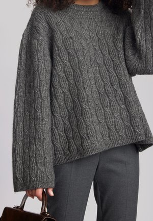 Person iført en grå kabelstrikket sweater og grå bukser, der holder en mørkebrun lædertaske i håndtaget.