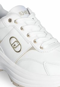Baskets blanches avec accents dorés, présentant une tige synthétique lisse, des lacets plats et une semelle légèrement surélevée et texturée. Logo sur le côté.