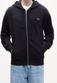Černá mikina na zip z bavlny s přední kapsou, kapucí se stahovací šňůrkou a logem "HUGO" na levé straně hrudi. Měkká textura, volný střih.