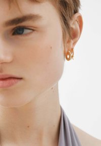 Pendientes de aro de oro con textura en espiral y acento de estrella. Primer plano de un rostro, con piel suave y ropa clara.