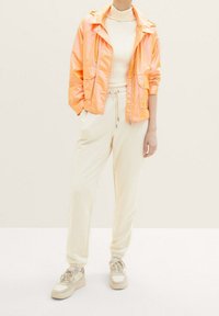 Giacca arancione brillante e lucida con cappuccio e due tasche, abbinata a pantaloni sportivi di tessuto crema e sneakers beige con accenti bianchi.