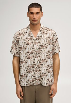 Junger Mann trägt ein kurzärmeliges Hemd mit beigefarbenen und braunen abstrakten Blatt- und Formmustern, kombiniert mit braunen Hosen.