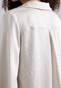 Camicia button-up di colore crema chiaro con un tessuto textured a pieghe, dotata di colletto classico e piega sul retro, che crea una silhouette rilassata.