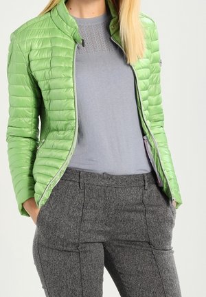 Kvinde iført en lysegrøn quiltet jakke over en grå strikket top og grå teksturerede bukser, med hænderne i lommerne.