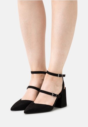 Chaussures en daim noir à talons carrés avec un bout pointu et deux brides à la cheville, dotées de boucles en argent et d'une texture lisse.