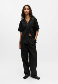 Schwarzes, gemustertes Kurzarm-Wickeltop mit V-Ausschnitt; passende lockere Hose; glattes Material mit dezentem Blätterdesign; getragen mit schwarzen Schuhen.