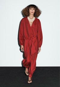 Rød linned jumpsuit med V-hals, lange ærmer og en knyttet bælte i taljen. Båret med et lagdelt perlehalskæde og sorte sandaler.
