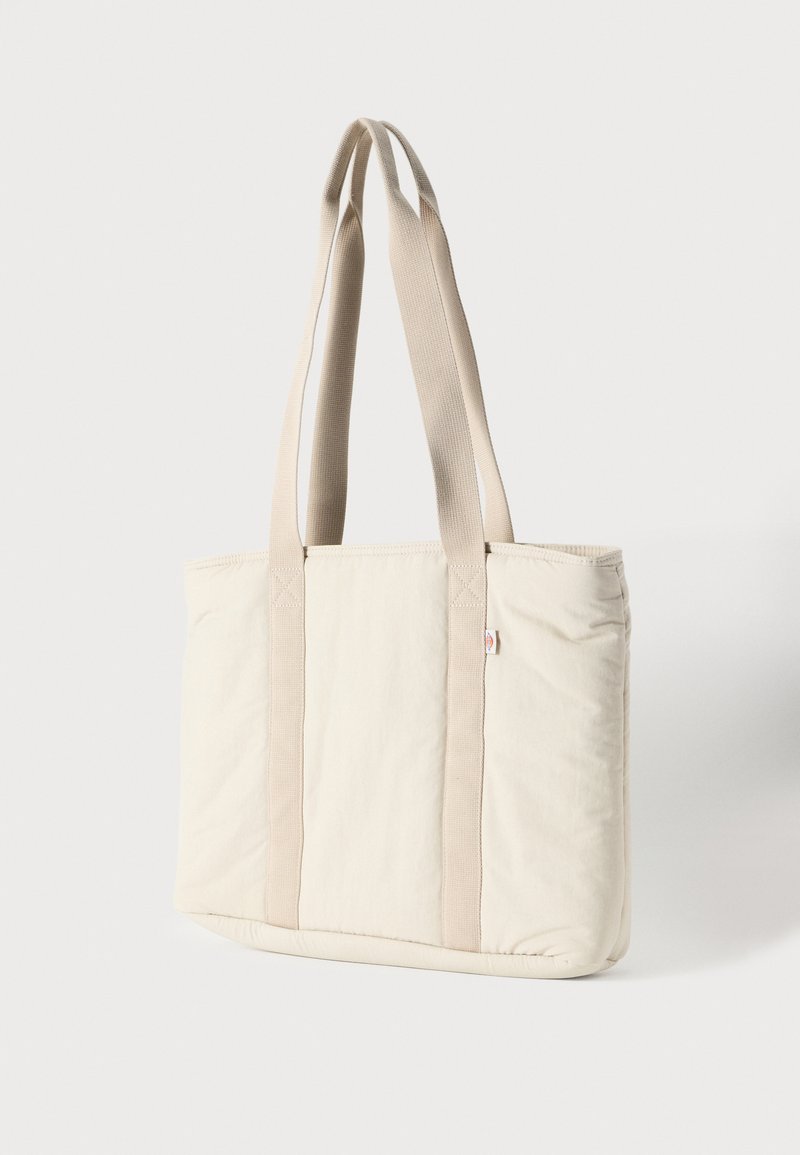 Borsa tote di tela beige con manici doppi lunghi e una piccola etichetta del marchio sulla parte superiore, posta su uno sfondo bianco uniforme.