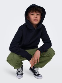 Felpa nera con cappuccio, pantaloni cargo verdi e sneakers alte nere. Tessuto morbido, vestibilità rilassata e polsini elastici su maniche e pantaloni.