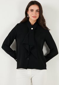 Black long-sleeve blouse with a collar and draped front. Features a shiny button closure and soft, textured fabric.