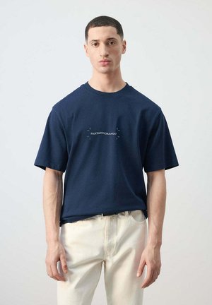 Joven vestido con una camiseta azul marino con texto "FANTASTIC MANGO" y pantalones blanco roto, de pie frente a un fondo claro y liso.